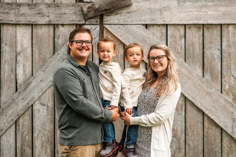 Smiling family of four posed in front of wooden barn – Imaginarium Pictures family photography