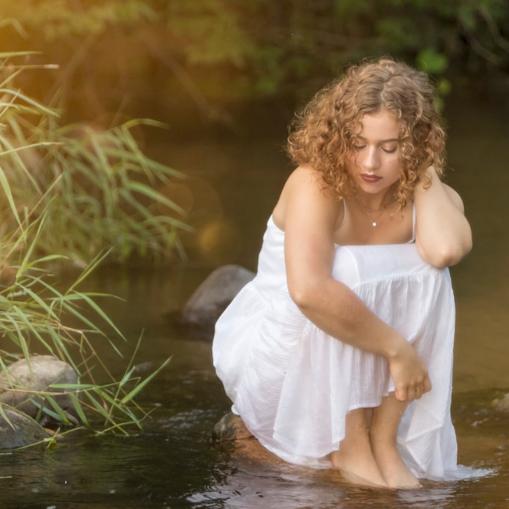 Natural senior portrait of a young woman with curly auburn hair sitting in a shallow stream, wearing a flowing white dress, soft golden-hour sunlight filtering through trees – senior photography by Imaginarium Pictures, Portage MI