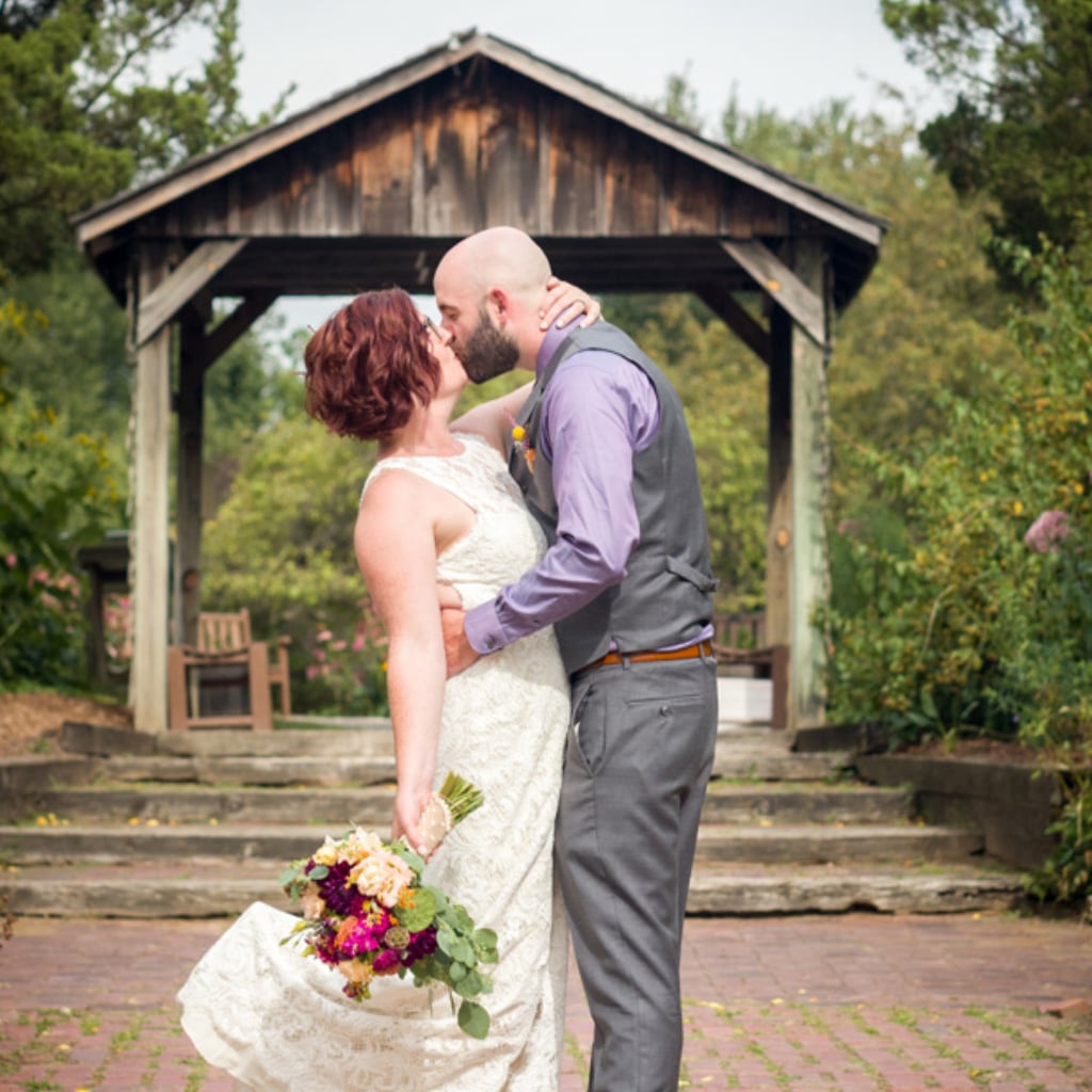 Bride with red hair in lace gown and groom in gray vest sharing a tender kiss under a rustic wooden gazebo, surrounded by lush green garden and blooming flowers – heartfelt wedding photography by Imaginarium Pictures, West Michigan