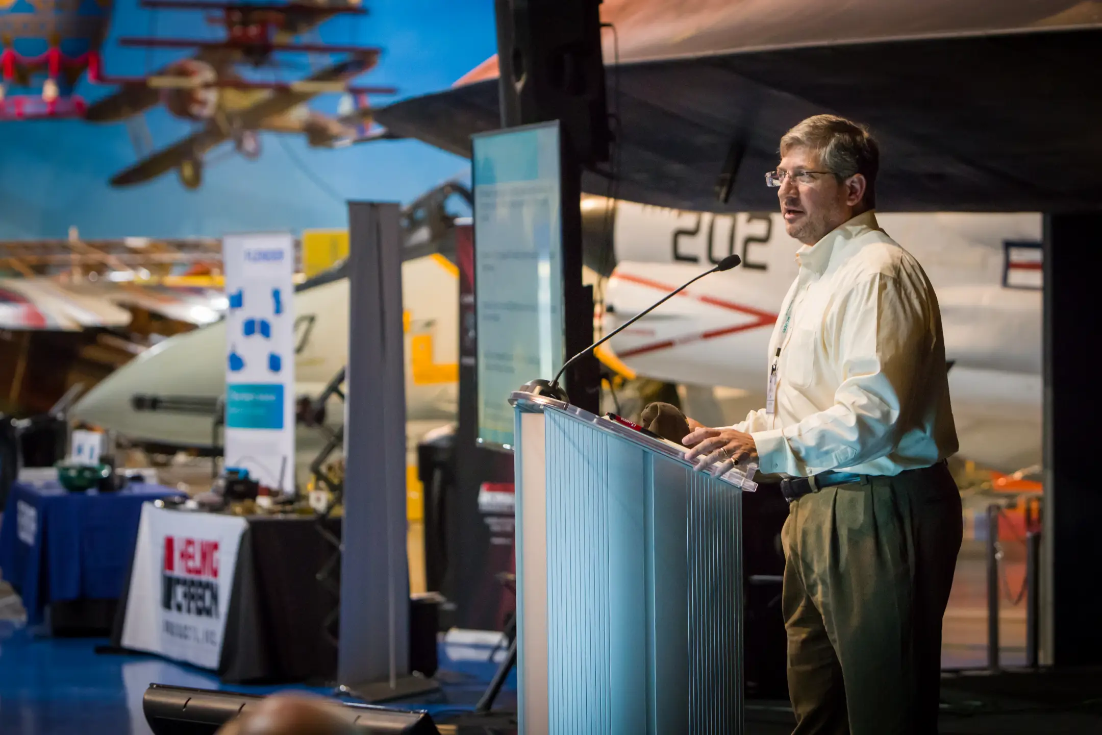 Natural senior portrait of a teenage girl with long wavy blonde hair in a white lace dress, leaning against a tree holding a bright sunflower, golden-hour string lights glowing in the wooded background – authentic senior photography by Imaginarium Pictures, West Michigan
